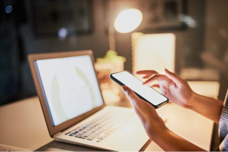 A desk with a laptop open and a pair of hands holding a cell phone.