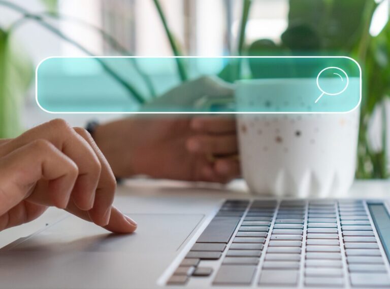 The hands of a woman are seen. One hand is holding a coffee cup, the other is scrolling on a laptop.