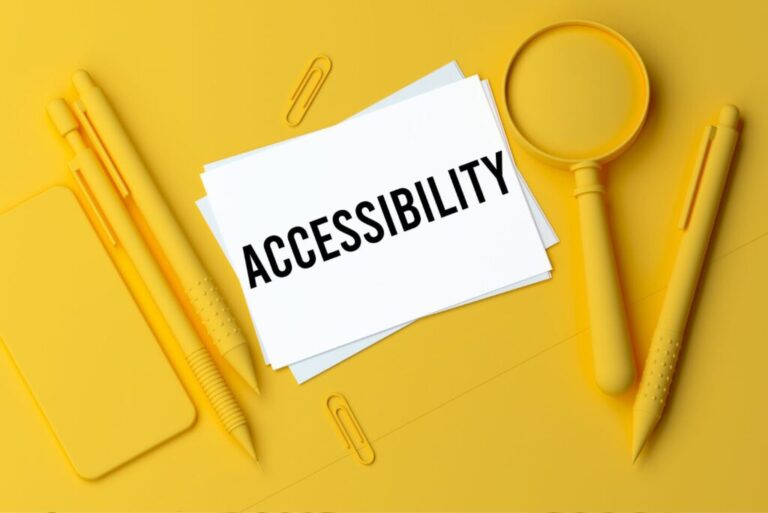 Yellow background with mobile phone, pens, and a magnifying glass. The word Accessibility is written on a white piece of paper.