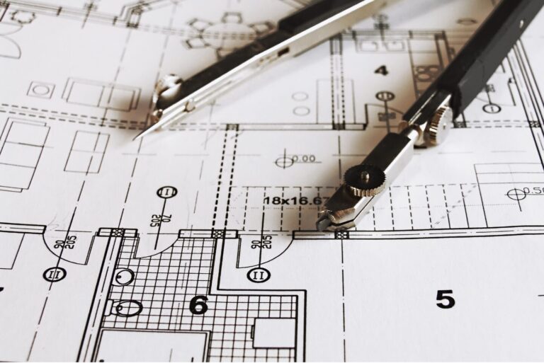 Architectual plans laid out on a table with a drawing compass on top.