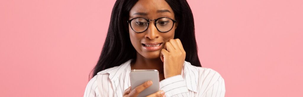 embarrassed woman cringing and looking at mobile phone with pink background