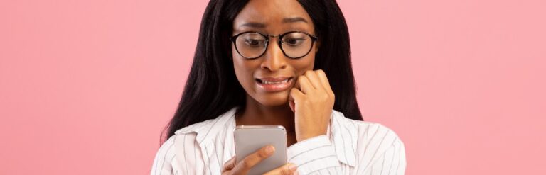 embarrassed woman cringing and looking at mobile phone with pink background