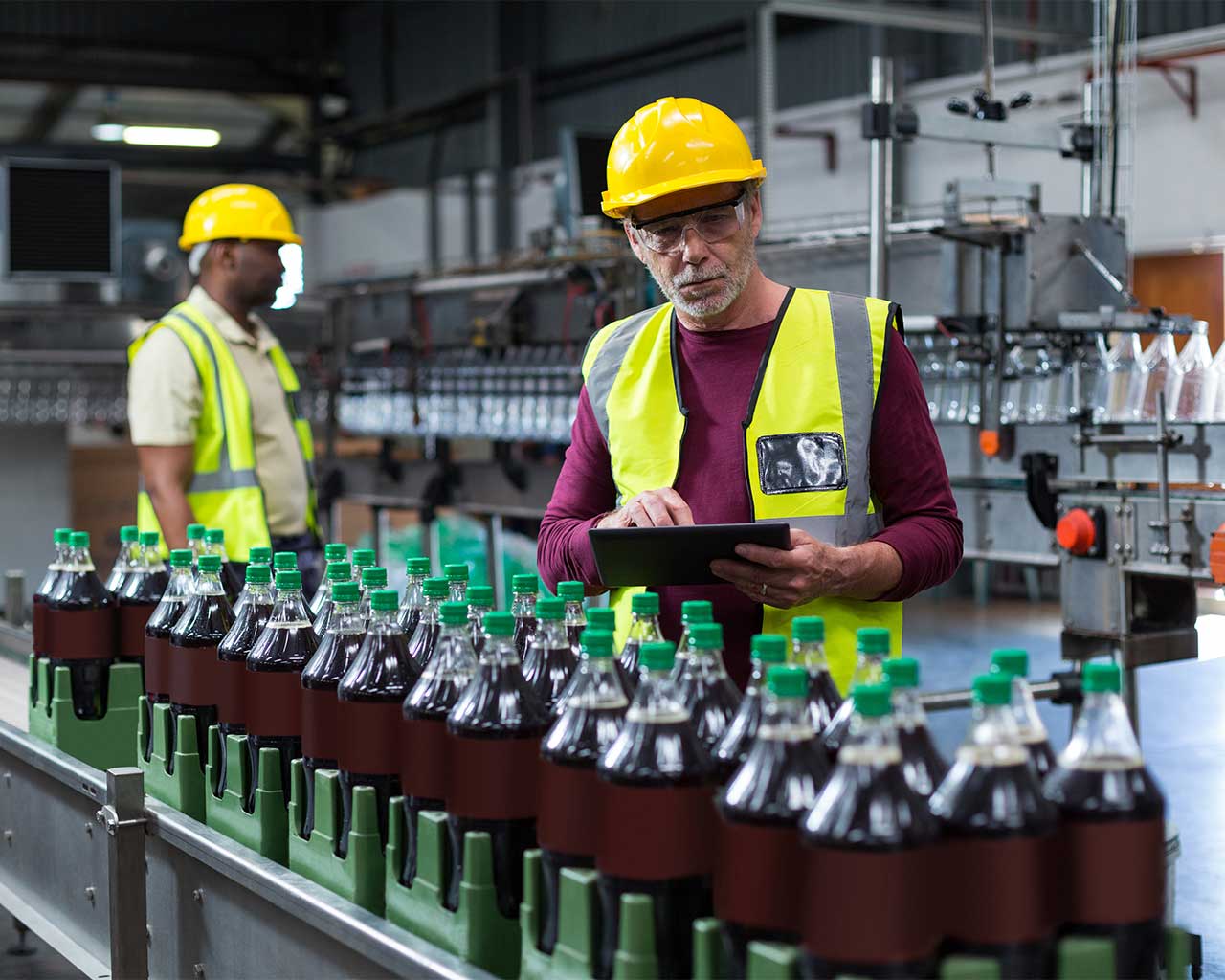 Factory workers wearing safety vests and hard hats inspect a beverage production line, with rows of bottled drinks moving along a conveyor belt.