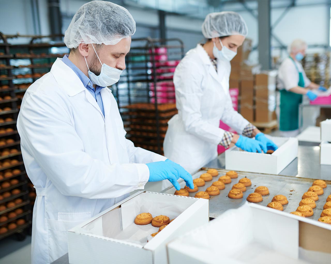 Food production workers in protective clothing and masks package baked goods on an assembly line inside a commercial bakery.