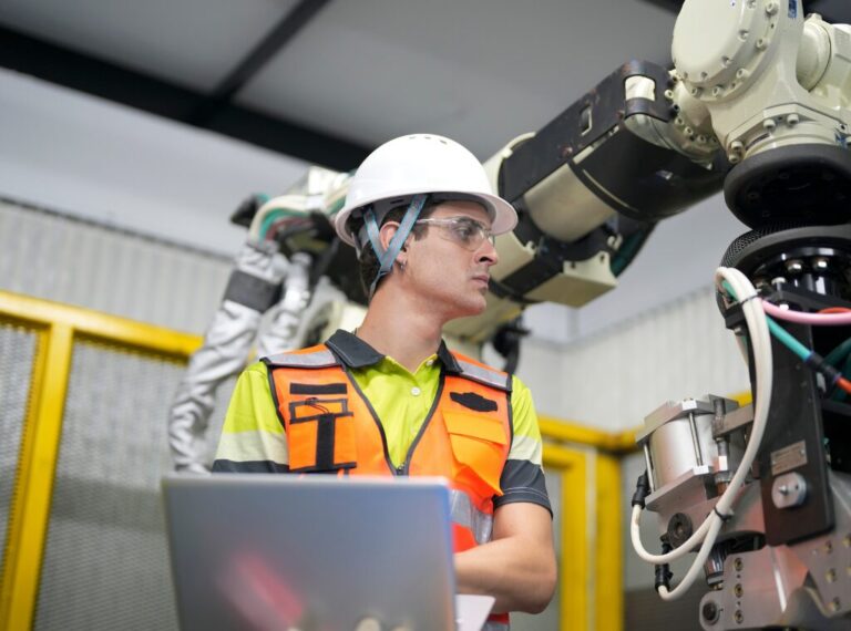 Man on his computer looking at a robotic arm in a manufacturing Wearhouse.