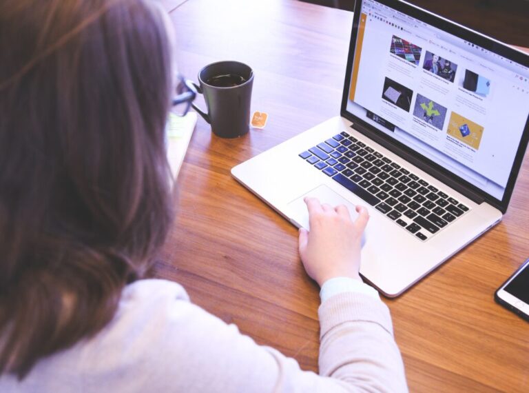 A woman is looking at her laptop. A cup of tea and cell phone are laying next to the laptop on the table. On the screen of the laptop is a homepage for a website.