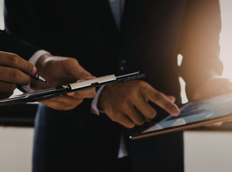 Two businessmen are looing at a tablet. One man is holding the tablet, while the other is taking notes on a notepad.